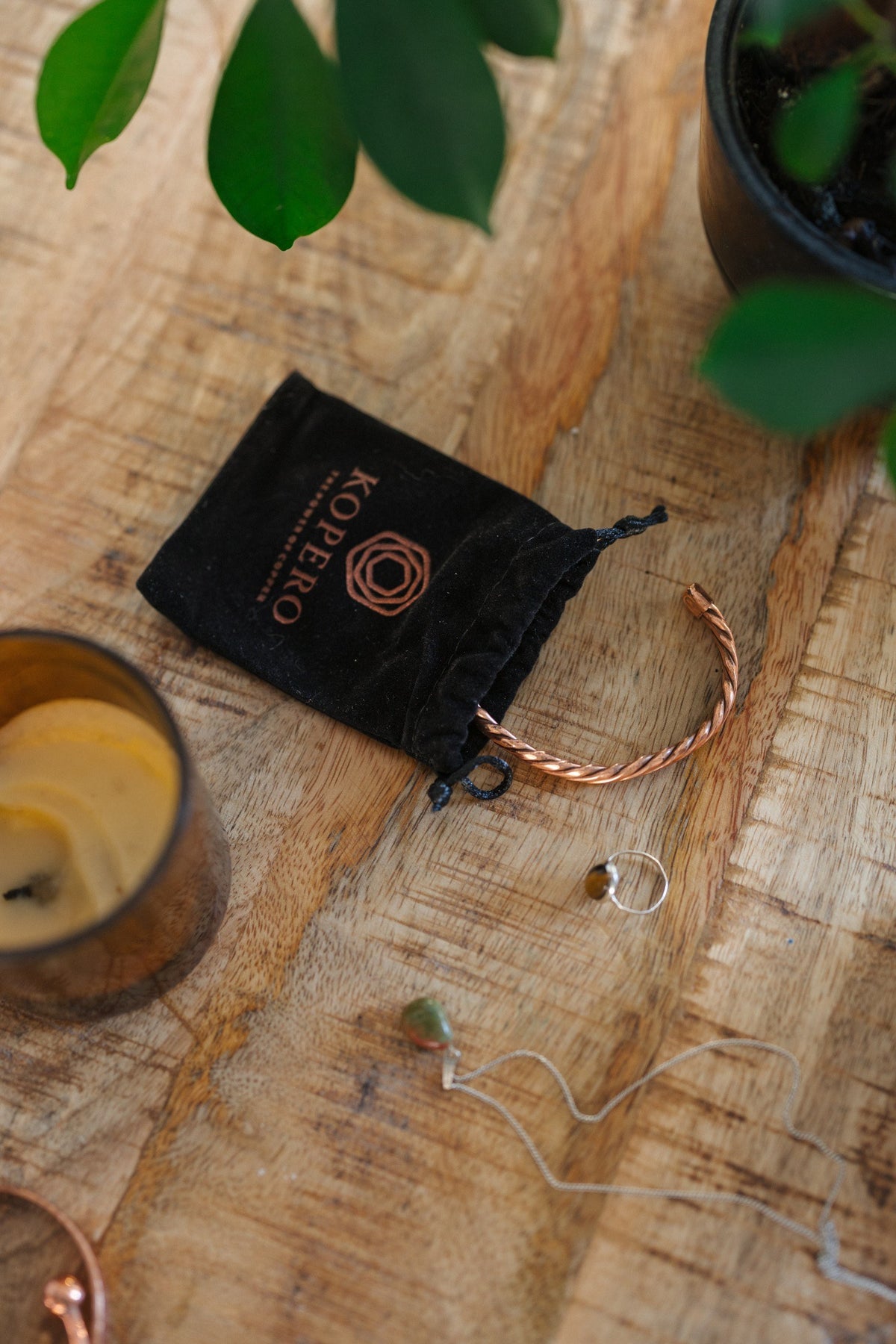 Kopero Copper Bracelet Twisted, on a wooden table next to the velvet pouch, a candle and jewellery