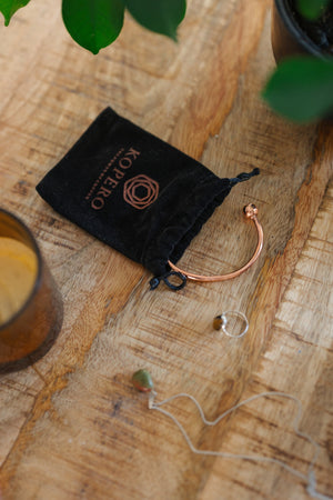 Kopero Copper Bracelet Round, on a wooden table next to the velvet pouch, a candle and jewellery