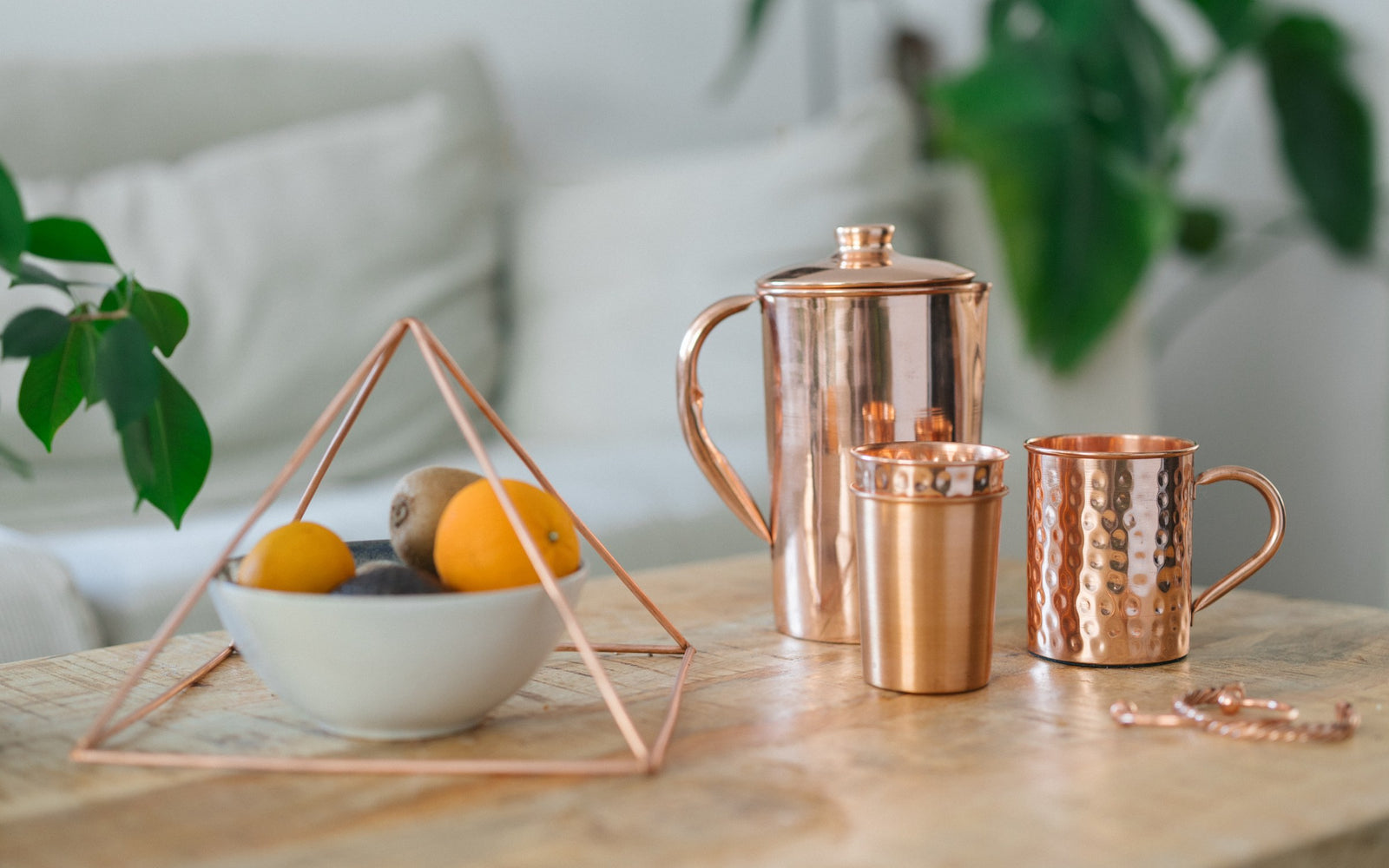 Copper pyramid over a bowl of fruit, with a jug, a cup, a mug and a bracelet on a wooden table