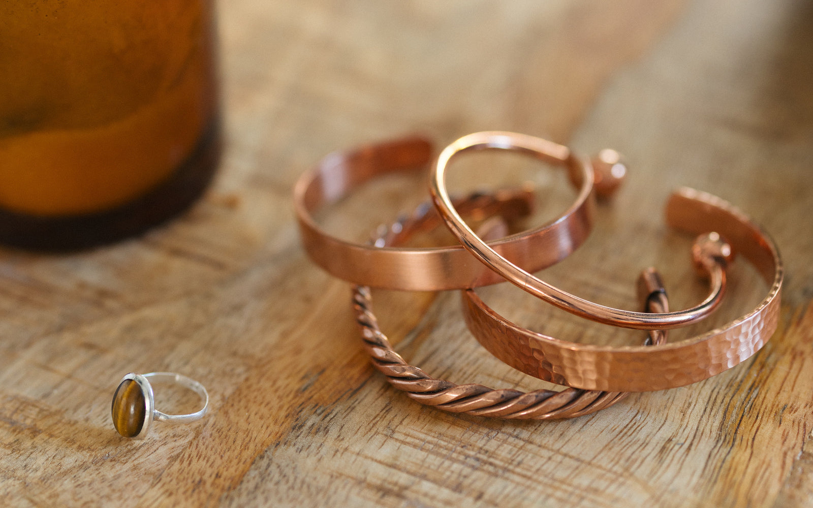 Four copper bracelets, twisted, smooth, hammered and round, on a wooden table next to a candle and a ring