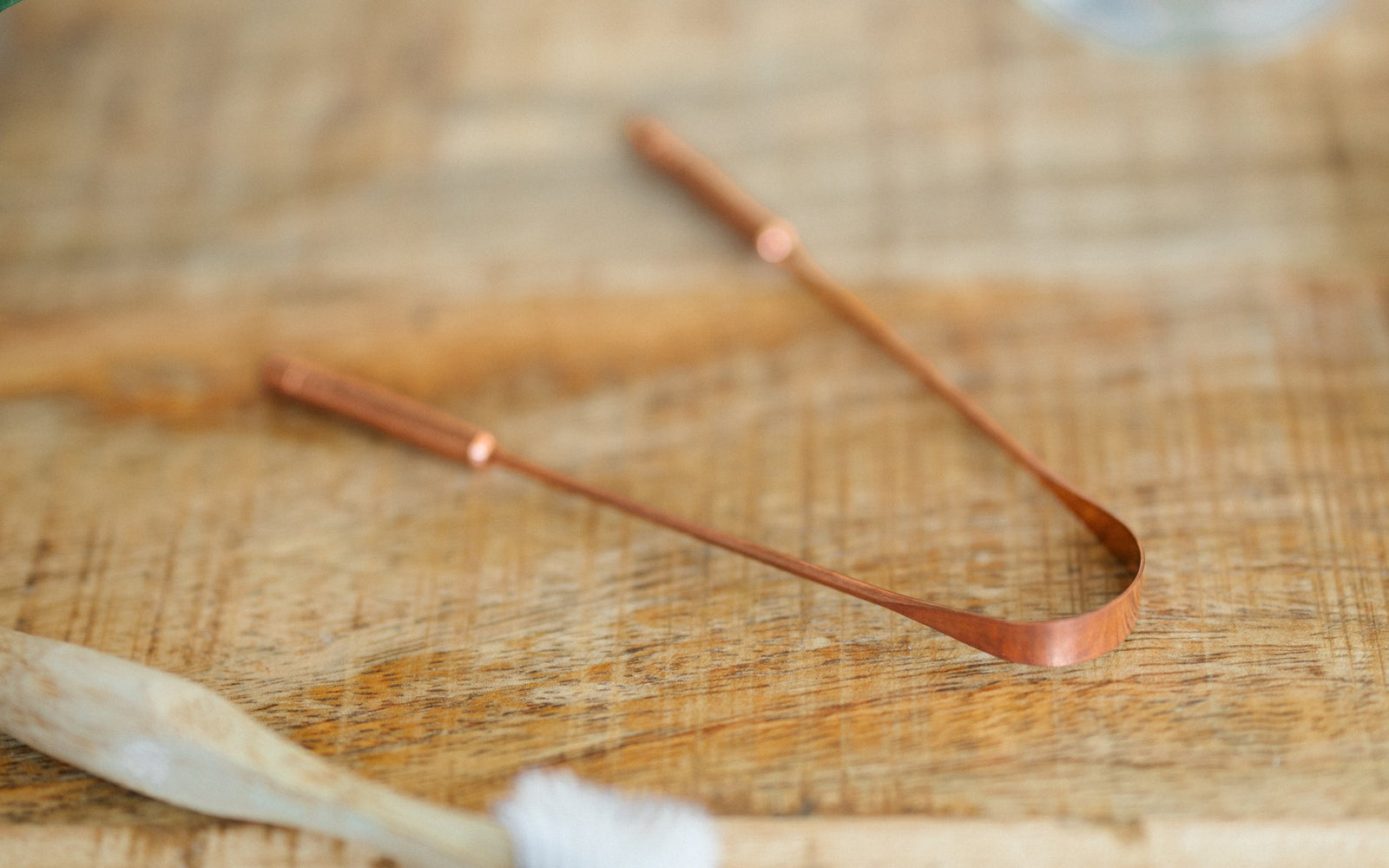 Copper tongue scraper on a wooden table next to a bamboo toothbrush