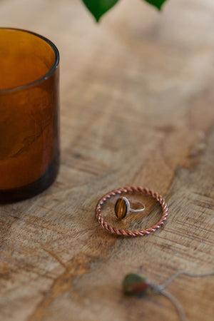 Kopero Copper Tensor Ring, on a wooden table with a tiger's eye ring and a glass candle holder