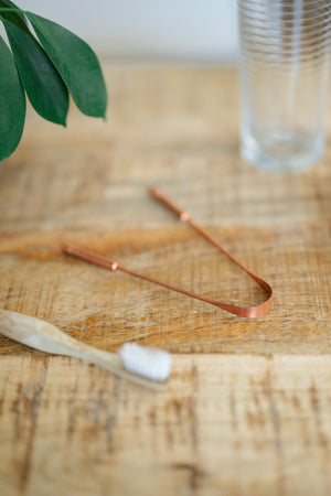 Kopero Copper Tongue Scraper, on a wooden table next to a bamboo toothbrush and a glass of water