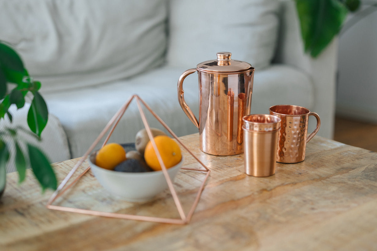 Copper pyramid over a bowl of fruit, with a jug, a cup and a mug on a wooden coffee table