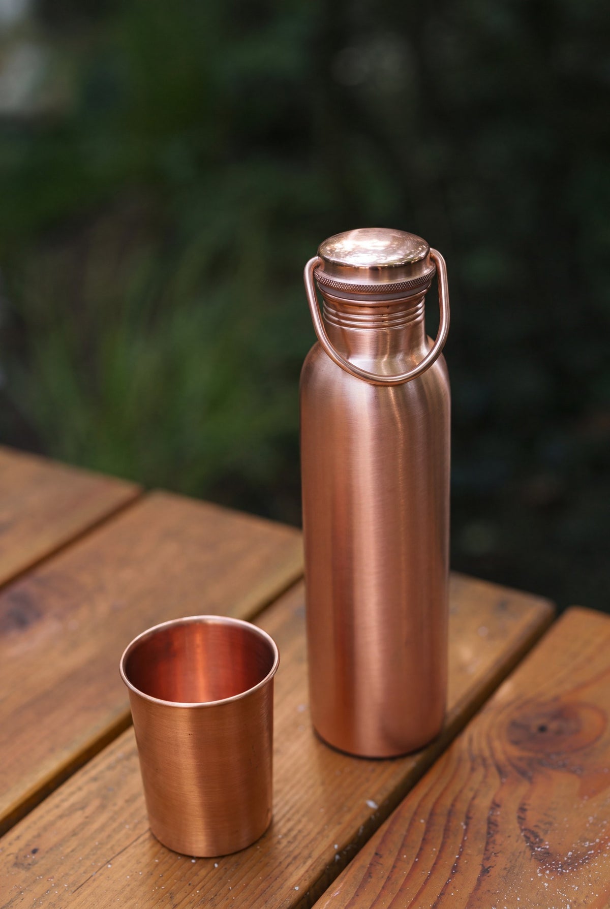 Kopero Copper Water Bottle Smooth, on a wooden garden table next to a copper cup